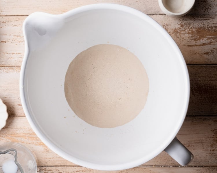 Overhead view of active dry yeast blooming in warm milk inside a white mixing bowl, showing foamy bubbles forming on the surface.