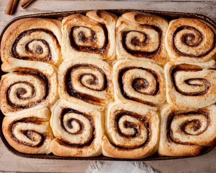 Overhead view of freshly baked cinnamon rolls in a baking dish, golden brown with soft centers and visible cinnamon swirls.