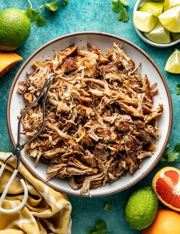 Pork carnitas in white serving bowl with brown rim with metal tongs on top.