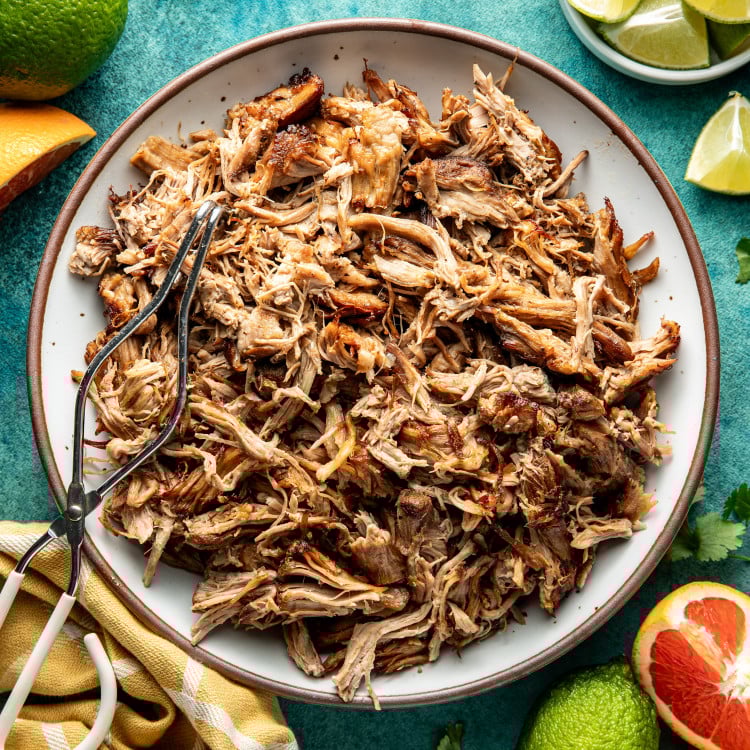 Pork carnitas in white serving bowl with brown rim with metal tongs on top.