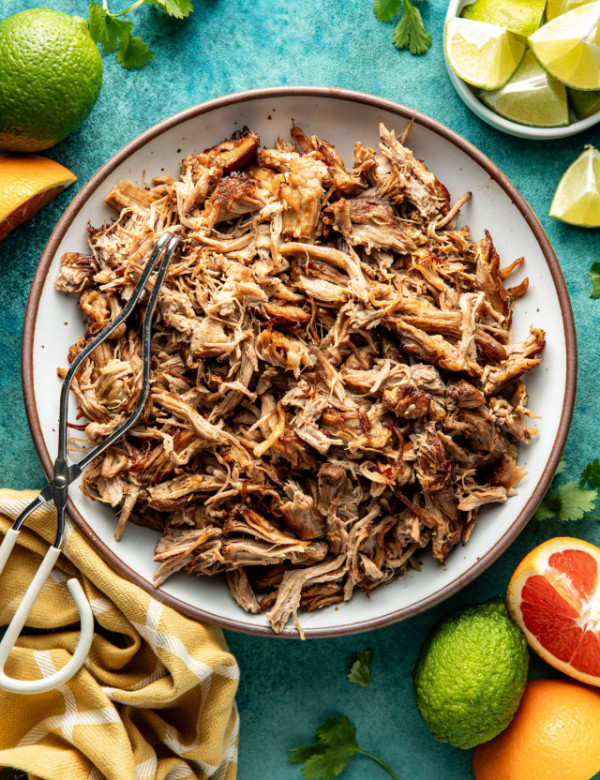 Pork carnitas in white serving bowl with brown rim with metal tongs on top.