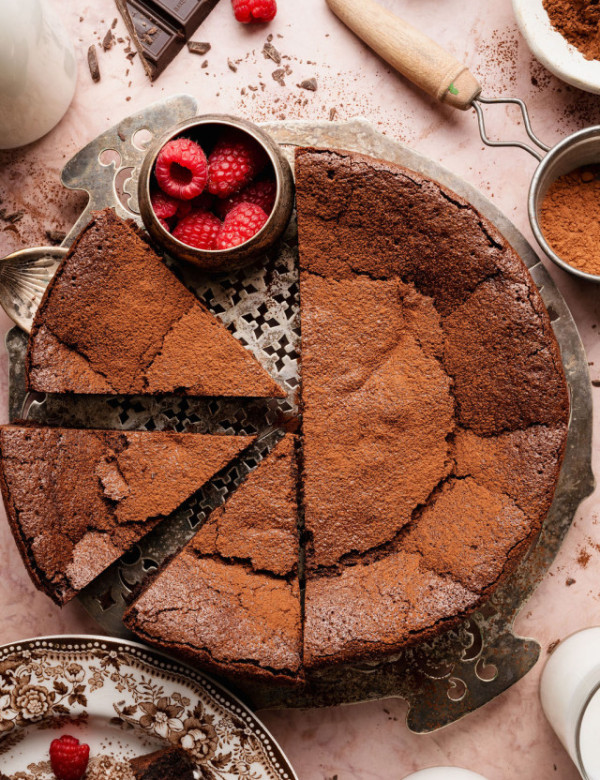 Overhead view of a flourless chocolate cake with multiple slices cut, garnished with cocoa powder and fresh raspberries on a metal cake stand.
