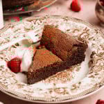 Slice of flourless chocolate cake dusted with cocoa powder on a decorative plate, served with whipped cream and a fresh raspberry, with the remaining cake visible in the background.
