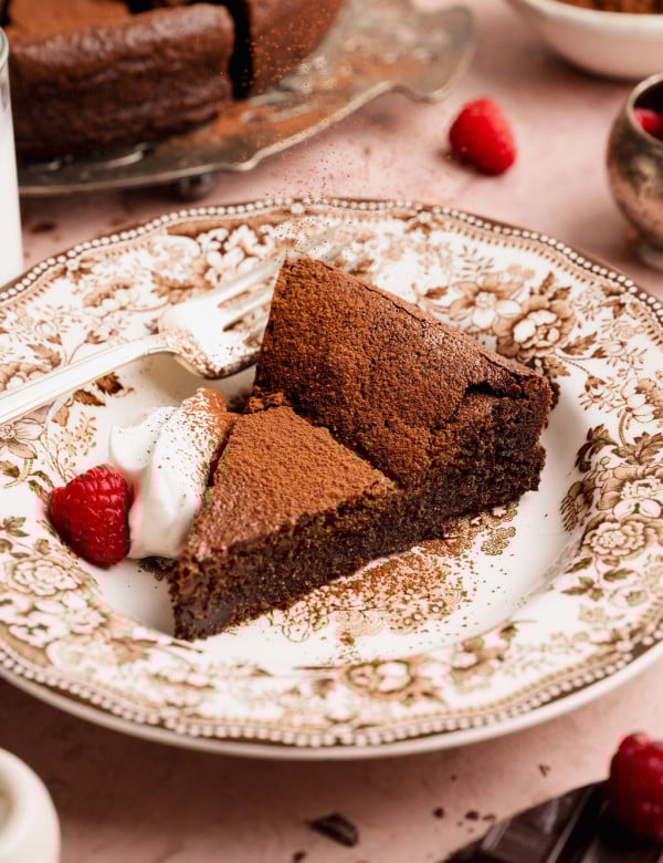 Slice of flourless chocolate cake dusted with cocoa powder on a decorative plate, served with whipped cream and a fresh raspberry, with the remaining cake visible in the background.