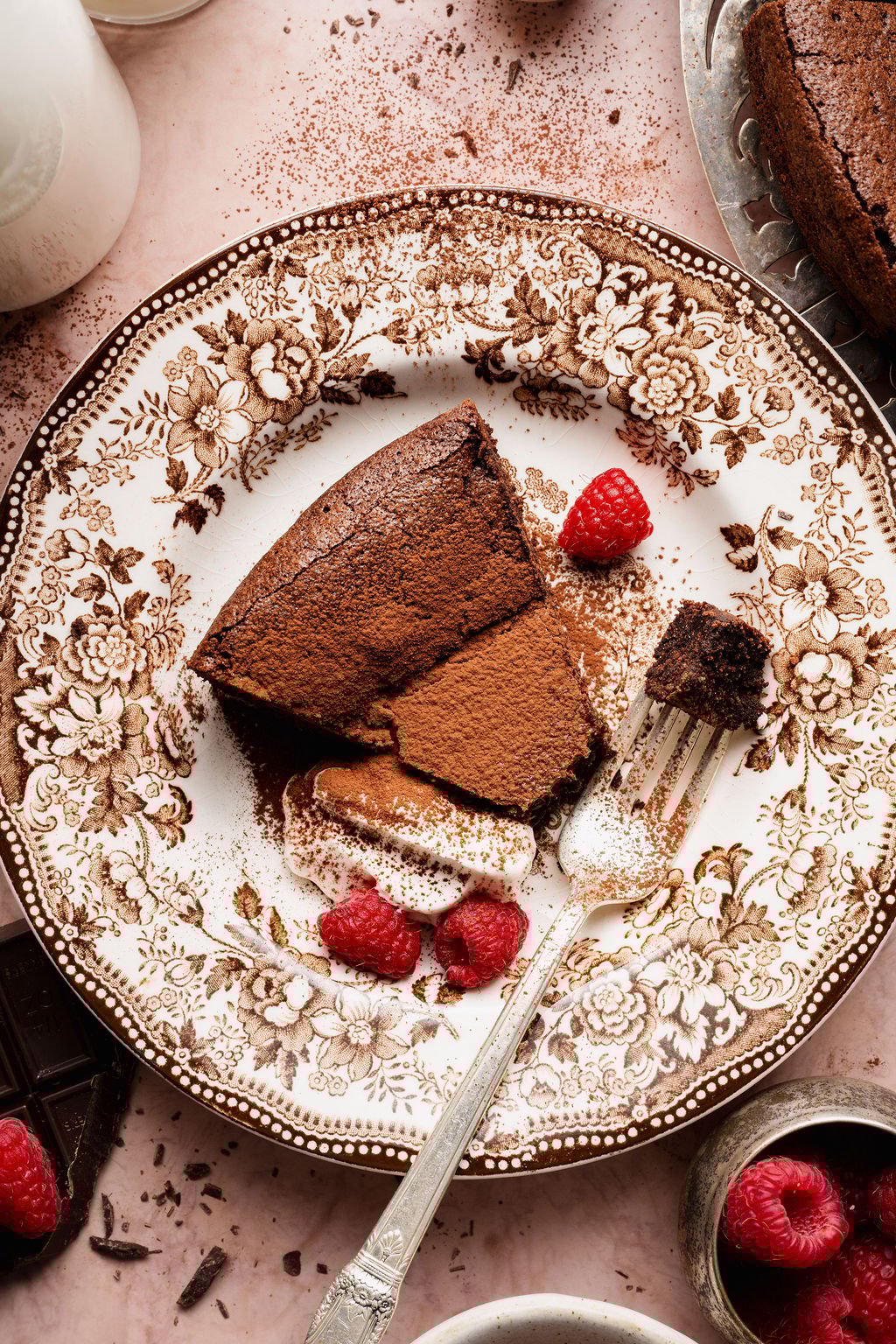 Slice of flourless chocolate cake dusted with cocoa powder, served with whipped cream and fresh raspberries on a vintage floral plate with a fork.