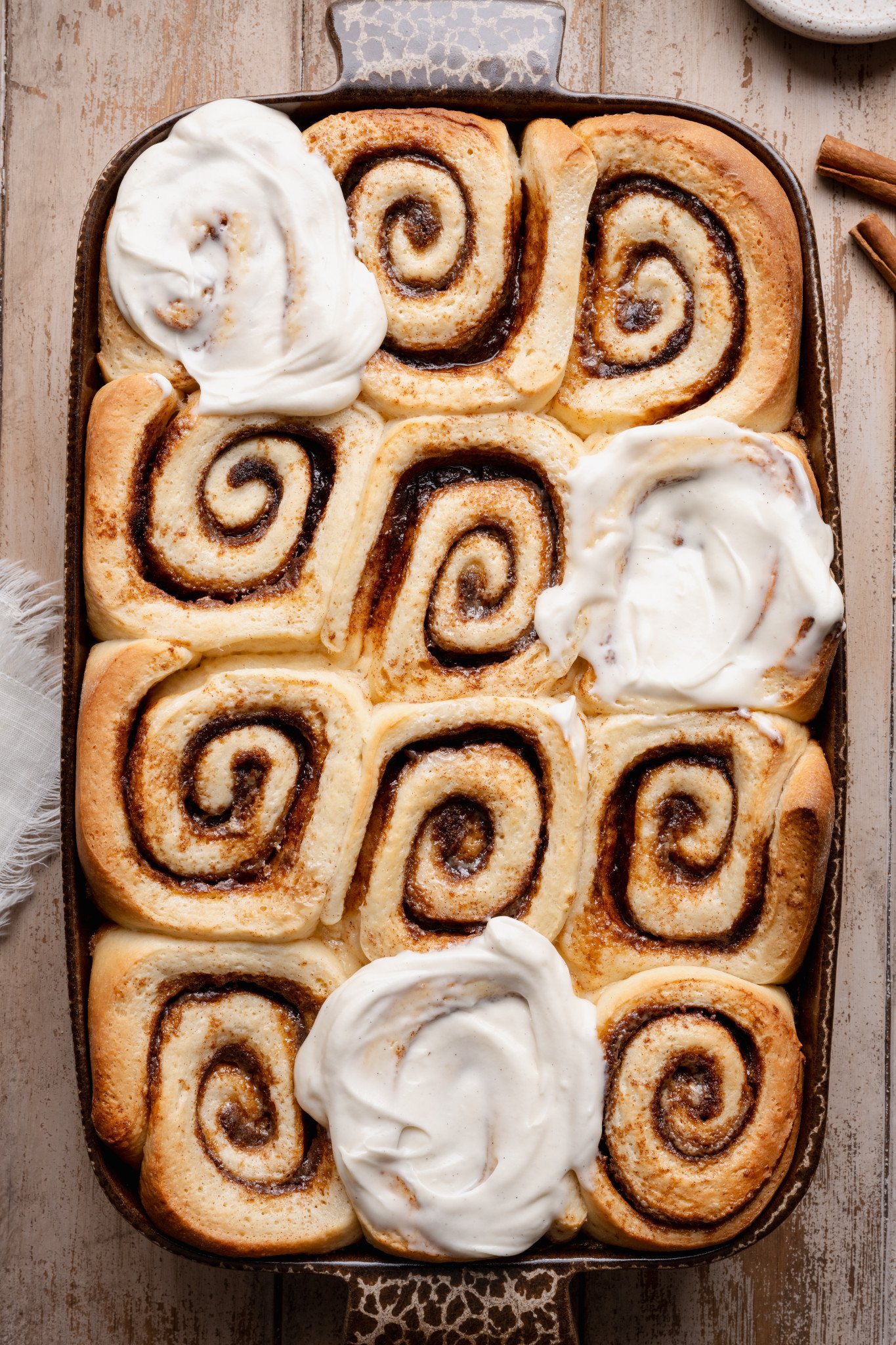 Overhead view of freshly baked cinnamon rolls in a ceramic baking dish with dollops of cream cheese frosting melting over the warm rolls.
