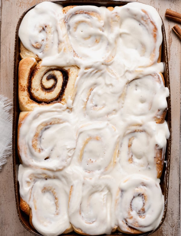 Overhead view of soft overnight cinnamon rolls baked in a ceramic baking dish and generously frosted with creamy vanilla cream cheese icing, showing fluffy swirls and golden edges.