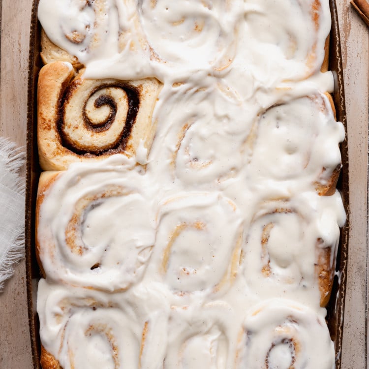 Overhead view of soft overnight cinnamon rolls baked in a ceramic baking dish and generously frosted with creamy vanilla cream cheese icing, showing fluffy swirls and golden edges.