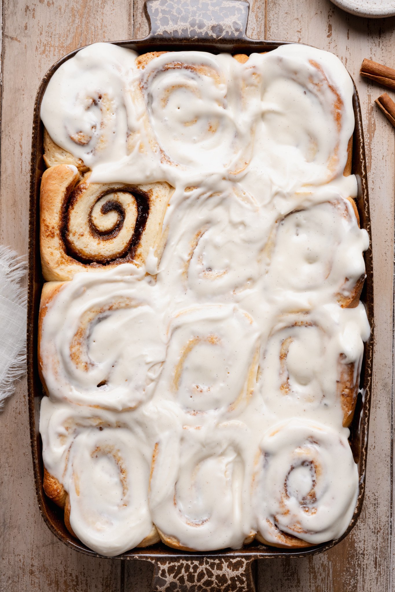Overhead view of soft overnight cinnamon rolls baked in a ceramic baking dish and generously frosted with creamy vanilla cream cheese icing, showing fluffy swirls and golden edges.