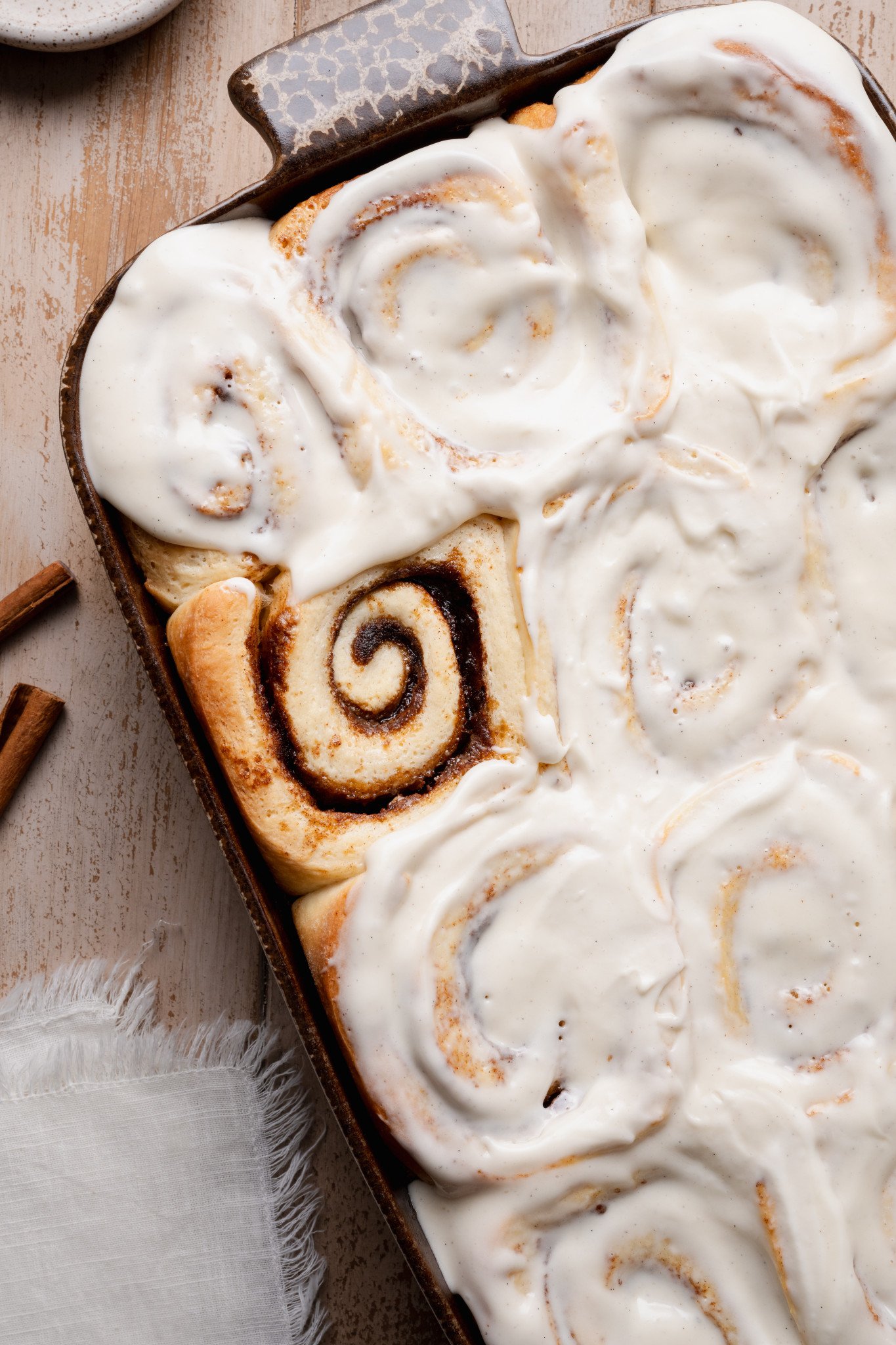 Close-up overhead view of cinnamon rolls generously frosted with creamy vanilla cream cheese icing in a rustic baking dish.