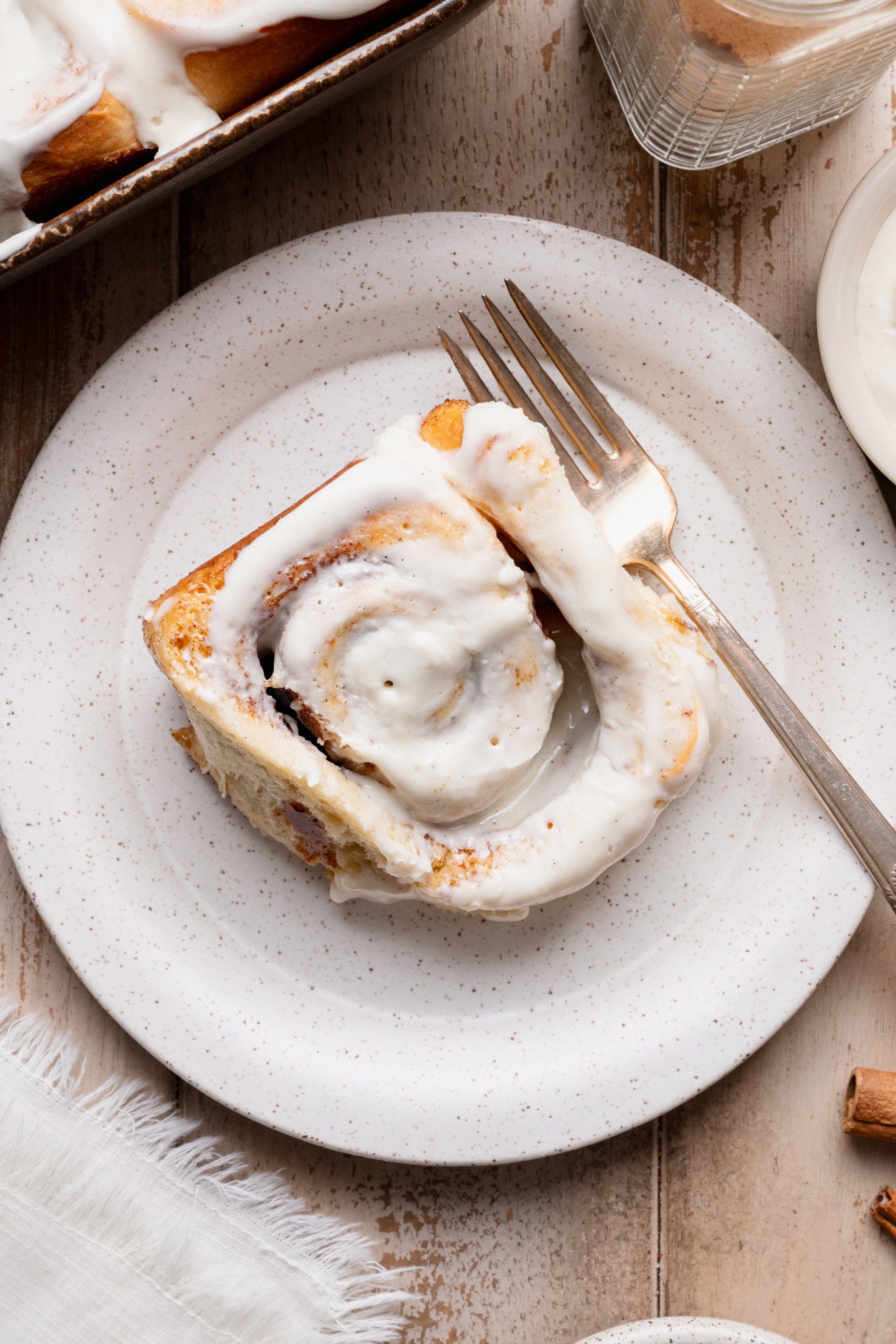 Plated cinnamon roll with cream cheese frosting and a fork, shown alongside a baking dish of frosted cinnamon rolls.