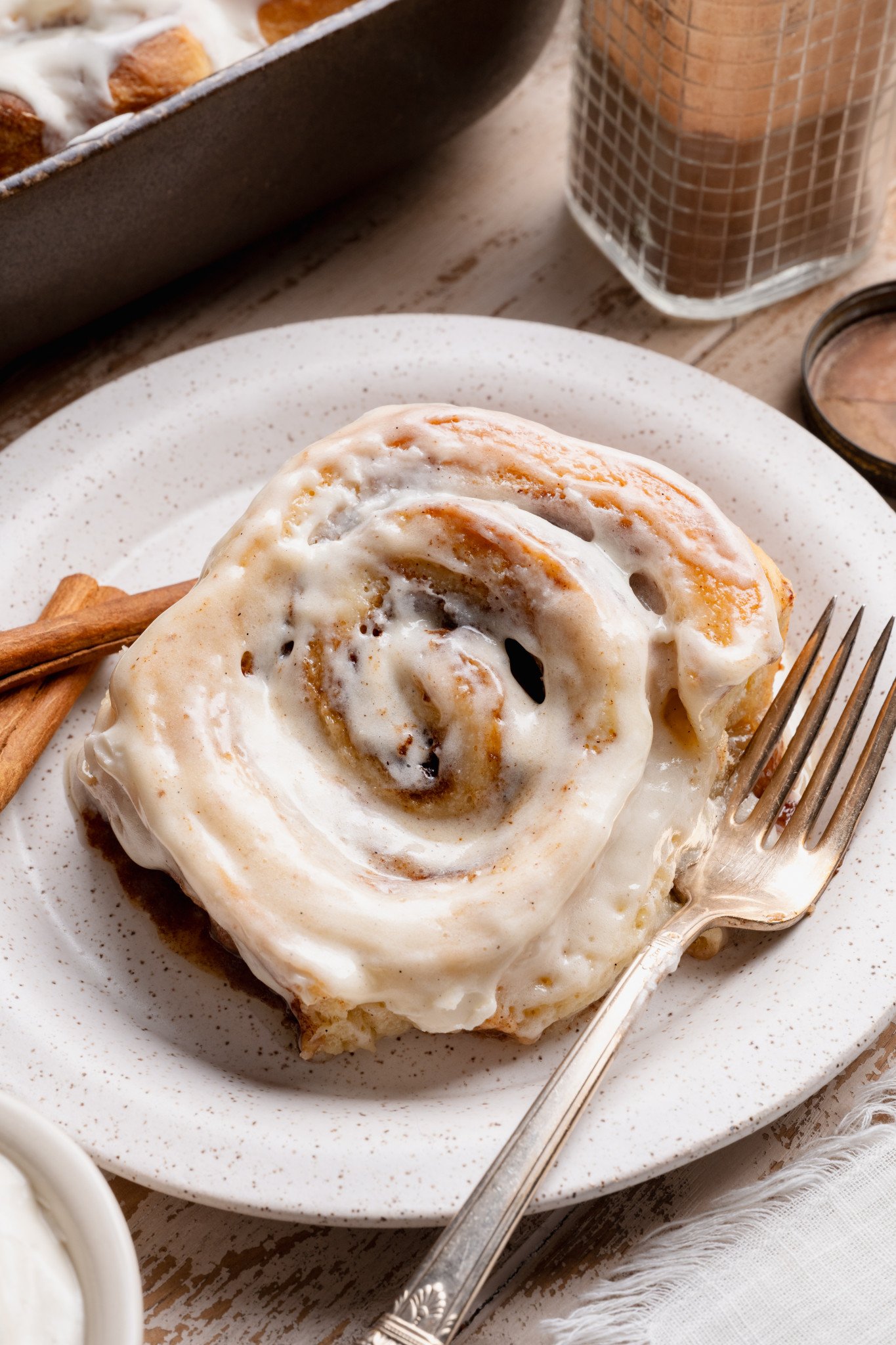 Close-up of a frosted cinnamon roll on a plate with cinnamon sticks nearby, highlighting the soft swirl and creamy frosting.