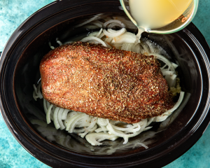 Assembling seasoned pork shoulder on top of onion and the rest of the ingredients for pork carnitas in slow cooker.