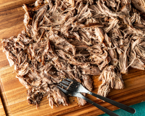 Cooked pork carnitas shredded with two forks on wood cutting board.