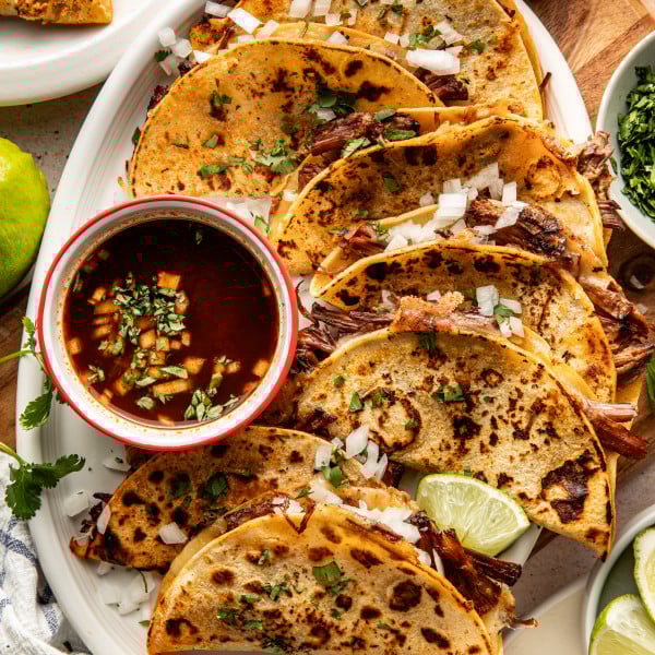 Birria tacos cooked and arranged on serving plate with bowl of consom&eacute; on side to dip.