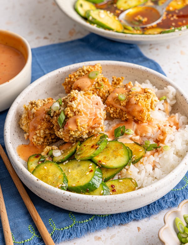 Salmon nuggets served in a bowl with rice and cucumber salad and drizzled with bang bang sauce.
