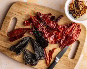 Dried guajillo chiles, ancho chiles, and arbol chiles arranged on a wooden cutting board with a knife, showing the different shapes and colors of the dried chiles before cooking.