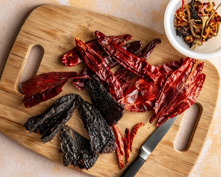 Dried guajillo chiles, ancho chiles, and arbol chiles arranged on a wooden cutting board with a knife, showing the different shapes and colors of the dried chiles before cooking.