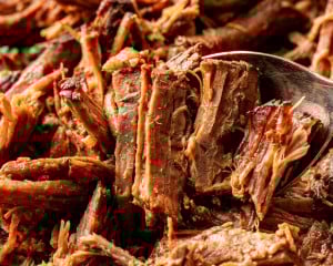 Close-up of shredded birria beef with a spoon lifting tender strands, highlighting the juicy texture and rich reddish-brown color.