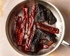 Dried guajillo chiles, ancho chiles, and arbol chiles toasted in a stainless steel skillet, highlighting their deep red and dark brown colors as they warm.