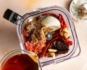 Birria sauce ingredients in a blender including softened dried chiles, roasted onion, garlic, diced tomatoes, spices, and broth being poured in.
