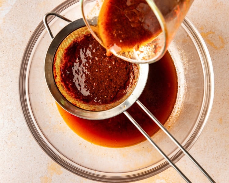 Birria sauce being strained through a fine mesh sieve into a bowl, separating smooth broth from chile solids.