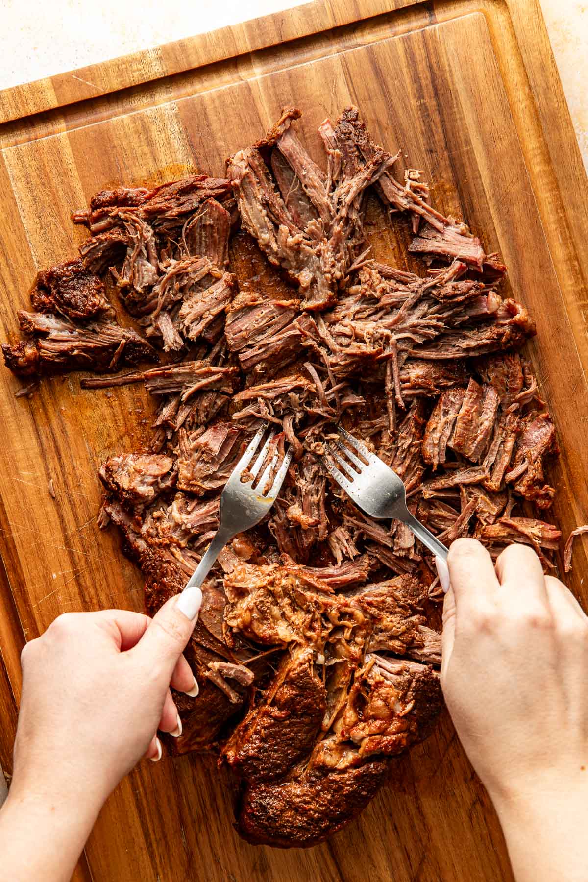 Hands using two forks to shred slow-cooked birria beef on a wooden cutting board, showing fall-apart texture and caramelized edges.