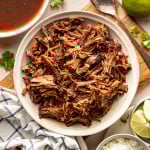 Overhead view of a bowl filled with shredded birria beef, with a bowl of reddish birria broth, lime wedges, cilantro, and diced onions arranged around it.