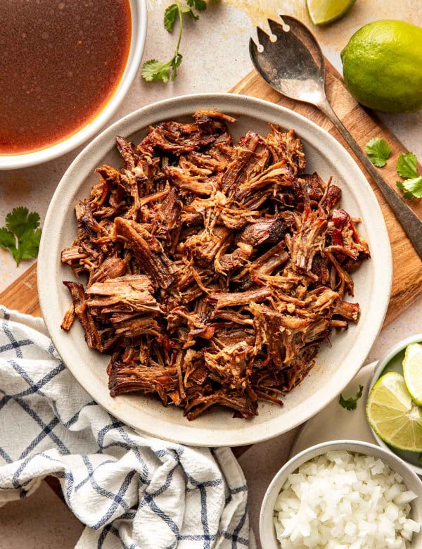 Overhead view of a bowl filled with shredded birria beef, with a bowl of reddish birria broth, lime wedges, cilantro, and diced onions arranged around it.