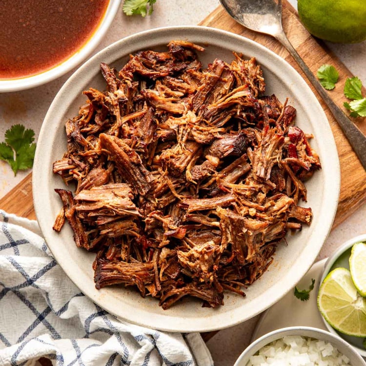 Overhead view of a bowl filled with shredded birria beef, with a bowl of reddish birria broth, lime wedges, cilantro, and diced onions arranged around it.