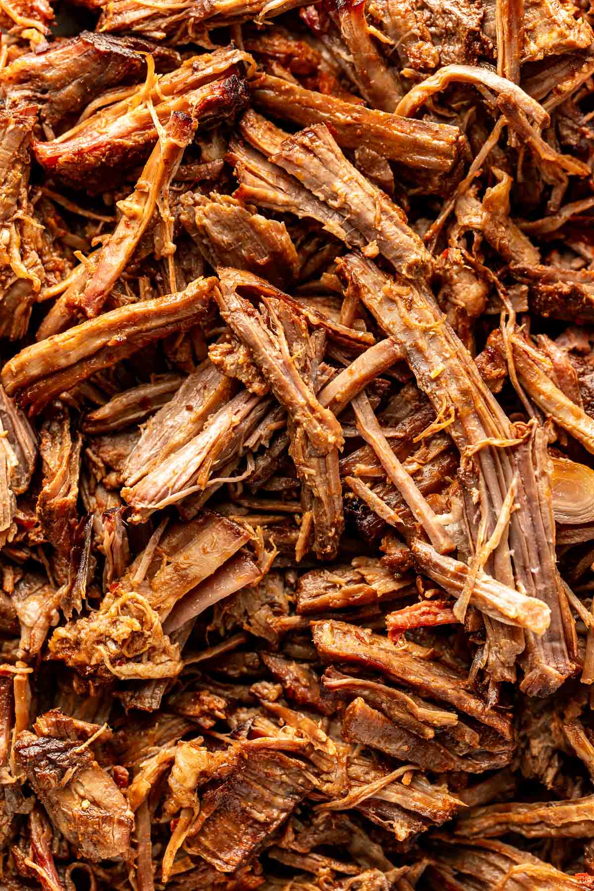 Extreme close-up of shredded birria beef filling the frame, showcasing tender fibers of beef coated in a rich, reddish-brown sauce.