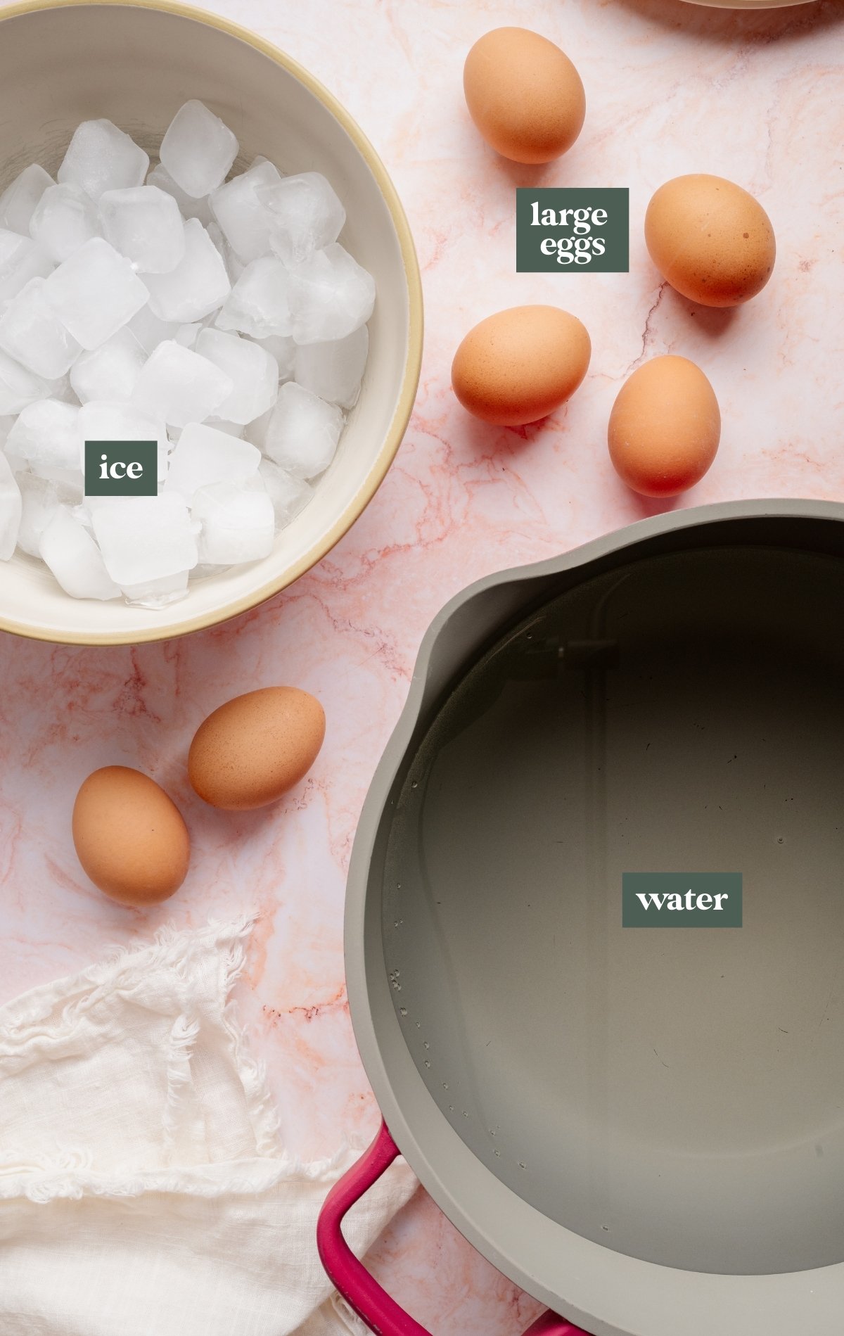 Overhead view of ingredients for stove top hard boiled eggs: a bowl filled with ice cubes, several large brown eggs, and a pot of water on a pink marble surface with a white linen towel nearby.