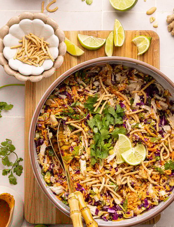 Thai chicken salad with peanut dressing in large bowl garnished with chow mein noodles and lime wedges.