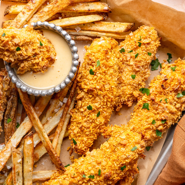 Tray of cornflake chicken tenders with fries and honey mustard dipping sauce with one tender being dunked into the dipping sauce.