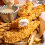 Cornflake chicken tenders on a tray with fries with one tender broken in half to show inside of chicken and one tender being dunked into the honey mustard dipping sauce.