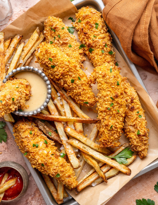 Baked cornflake chicken tenders on tray with fries and one tender being dunked into dipping sauce.