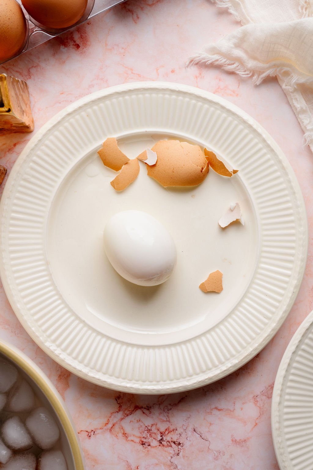 Peeled hard boiled egg on a white plate with cracked brown eggshell pieces scattered around on a pink marble surface.