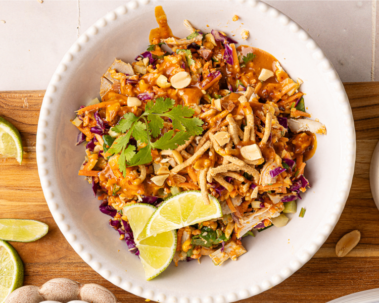 Thai chicken salad served in a small scalloped white bowl topped with chow mein noodles, dressing, cilantro and lime wedges.