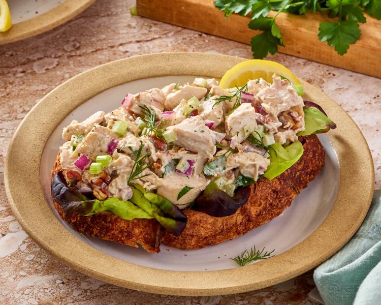 Chicken salad on top of bread garnished with lemon slice and fresh herbs on a small plate.