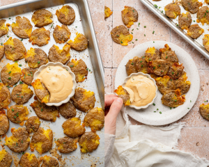 Side-by-side image of baked crispy smashed potatoes on baking sheet then plated and being dipped into small bowl of sauce.