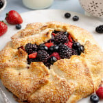 Golden, rustic mixed berry galette with a flaky, sugar-sprinkled crust folded around a juicy filling of blackberries, blueberries, and sliced strawberries, served on a white plate. Fresh berries are scattered around the plate, with a small colander of berries and bottles of milk softly blurred in the background, creating a bright, fresh kitchen scene.