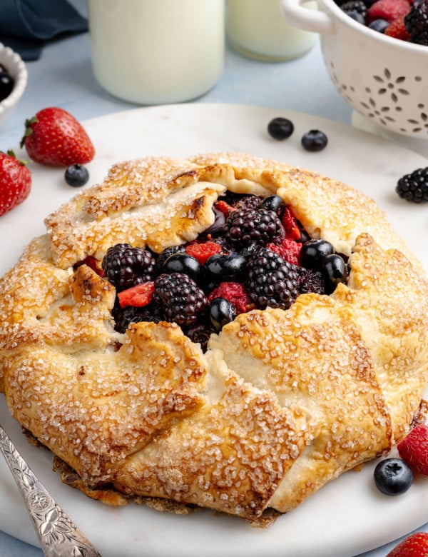 Golden, rustic mixed berry galette with a flaky, sugar-sprinkled crust folded around a juicy filling of blackberries, blueberries, and sliced strawberries, served on a white plate. Fresh berries are scattered around the plate, with a small colander of berries and bottles of milk softly blurred in the background, creating a bright, fresh kitchen scene.