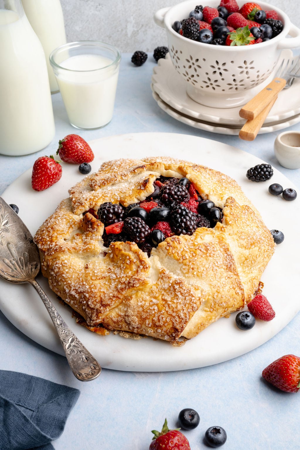 Angled view of a rustic mixed berry galette with a crisp, sugar-dusted crust and juicy berry filling, surrounded by fresh berries and a glass of milk on a light background.