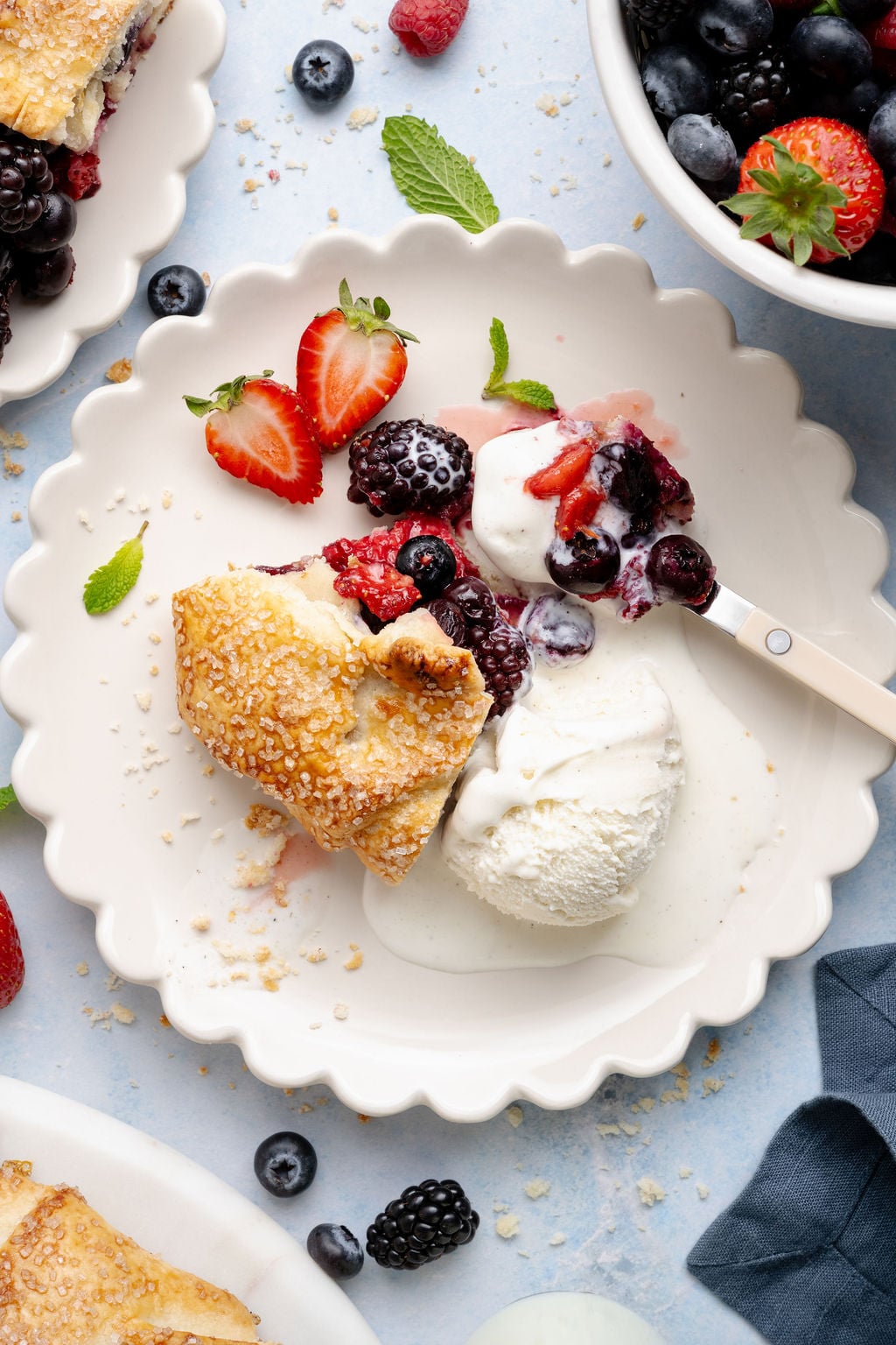 Overhead view of a plated slice of mixed berry galette served with a scoop of vanilla ice cream, fresh berries, and mint leaves on a scalloped plate.