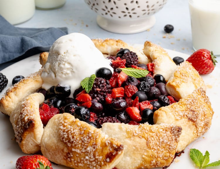 Close-up angled view of a baked mixed berry galette with a scoop of melting vanilla ice cream, highlighting the golden, flaky crust and vibrant berry filling.