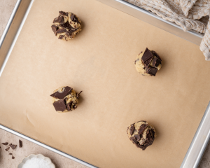 Four chocolate chunk cookie dough balls on a baking sheet with parchment paper.