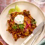 Chicken and rice enchilada casserole on a plate with a fork and knife garnished with pickled red onions, jalapenos, sour cream and cilantro.