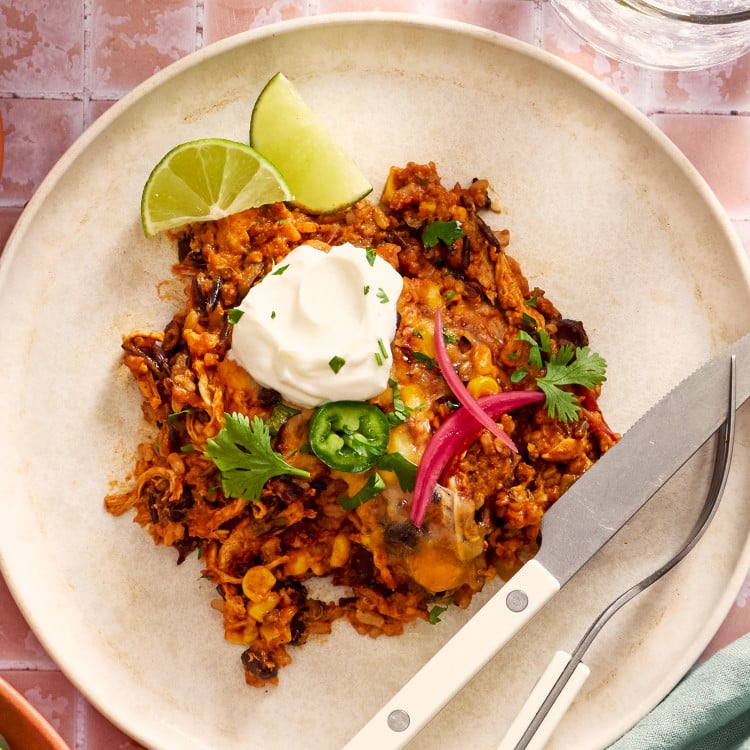 Chicken and rice enchilada casserole on a plate with a fork and knife garnished with pickled red onions, jalapenos, sour cream and cilantro.