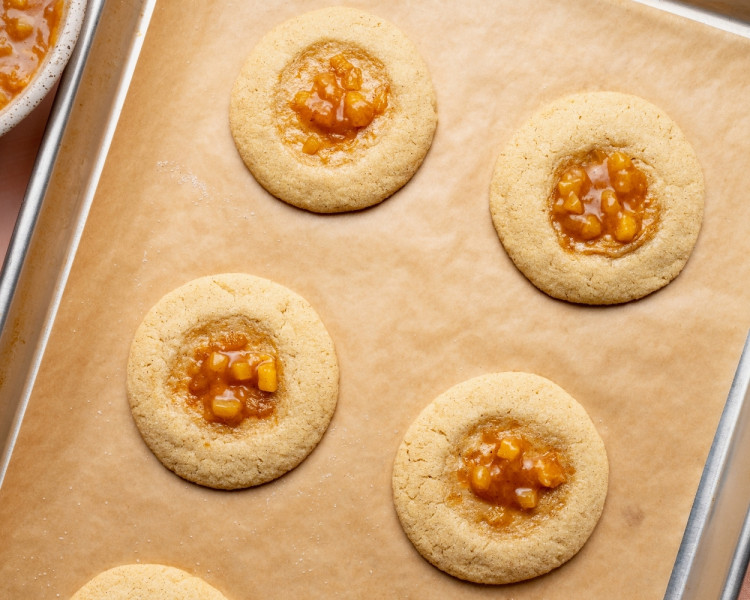 Overhead image of baked peach cobbler cookies on a parchment-lined baking sheet, each cookie filled with glossy homemade peach cobbler filling in the center wells before glazing.