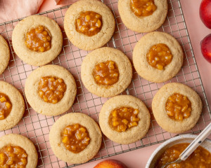 Overhead image of peach cobbler cookies cooling on a wire rack with homemade peach filling spooned into the centers. Fresh peaches and a bowl of peach filling are styled around the cookies on a blush pink background.